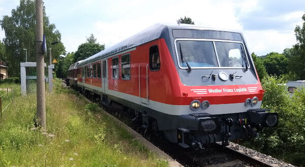 Roter ÖPNV Zug fährt auf Gleisen durch grüne Landschaft mit Bäumen und Stromleitungen.